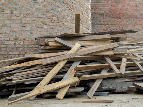 A collection of discarded wooden planks and beams is stacked haphazardly next to a brick wall at a construction site during the day