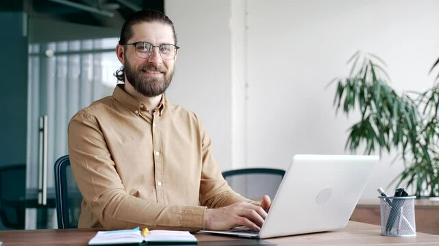Portrait of smiling IT specialist businessman sitting at desk at workplace in modern business office. Successful handsome coder developer busy with project working on laptop looking at camera