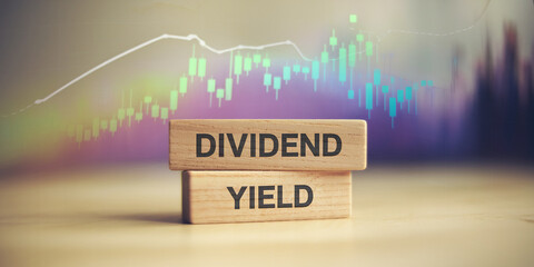 Wooden blocks display "Dividend Yield" against a blurred stock market graph background, symbolizing investment return and financial growth