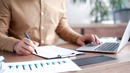 Close up of a male hand filling out documents with a pen using a laptop computer at a desk in an office. The businessman is engaged in paperwork. A financier or accountant fills out a blank tax form