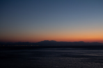 A vibrant sunset over the sea in Loutraki, Greece. Warm golden and pink tones fill the sky above the calm water, creating a romantic and tranquil moment.