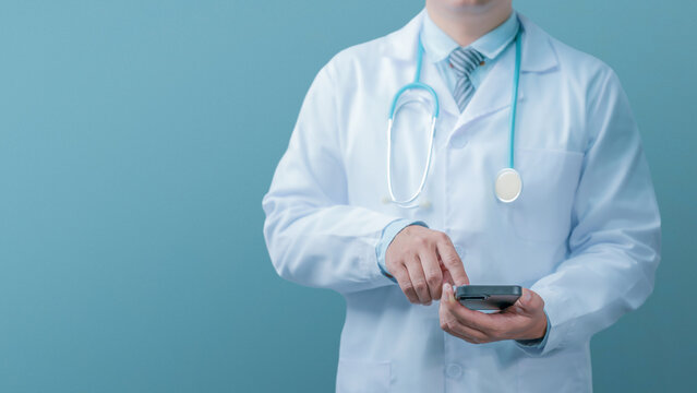 Medical technology and healthcare concept. A doctor in a white lab coat and stethoscope is using a smartphone against a plain blue-grey background. Online medical records.