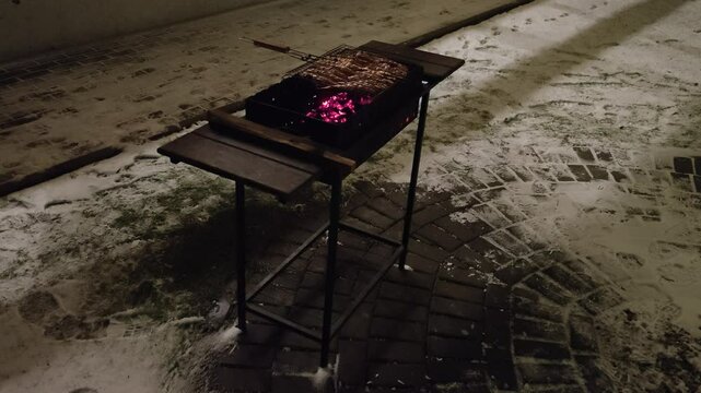 Winter evening twilight. In the yard, a metal brazier stands on a tiled area next to a lawn and the wall of the house. Coals are smoldering under a metal grate with wooden handles filled with mullet f