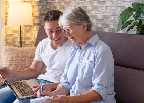 Young adult sitting on couch at home helps his elderly grandmother surf the web on laptop while she takes notes. The younger generation caring for older relatives by teaching them new technologies