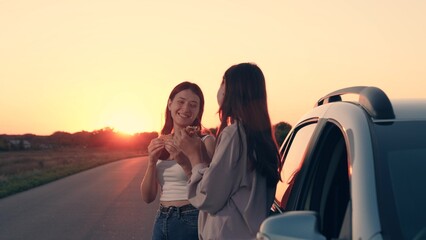 beautiful young girls eating sandwiches standing by car rays sunset, road food, long trip,...