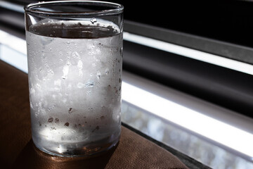 A cold glass of water with condensation sits on a brown surface by the window, catching natural light. The droplets suggest freshness and coolness, ideal for hydration or lifestyle themes.