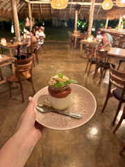 Hand holding a dessert in a jar topped with fruit sauce and mint, served on a plate with a spoon, in a cozy tropical restaurant with warm lighting and wooden furniture.
