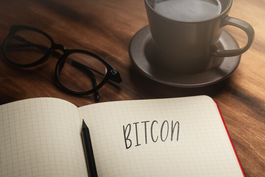A handwritten inscription "Bitcoin" on a grille of an open notebook on a wooden countertop, next to a black pencil, a cup with coffee and glasses, a flash of light. (selective focus)