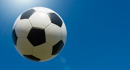 Naklejka premium A black and white soccer ball is shown in front of a clear blue sky with intentional lens flare. It represents sport and game, suitable for championship content.