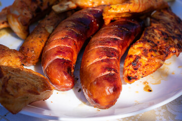 Close-up of grilled meat skewers and sausages on a white plate, served outdoors on a sunlit table, perfect for a summer barbecue.
