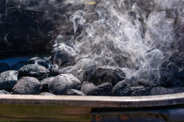 Close-up of charcoal briquettes heating in a grill with white smoke rising. The coals are still mostly black, indicating the early stage of lighting.