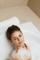 Relaxed young woman enjoying a bubble bath in a modern bathroom, with soft natural light enhancing her peaceful expression. Self-care and wellness concept.
