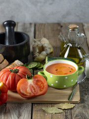 Close-up of a nice plate of cold gazpacho along with several tomatoes, toasted bread and various ingredients on a wooden table
