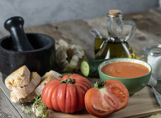 Green bowl of tomato gazpacho along with various ingredients and kitchen utensils on a wooden table