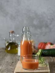 A vertical photo of a glass of gazpacho and a bottle in the background, on a tray isolated on a wooden background, with ingredients in the background. Copy space