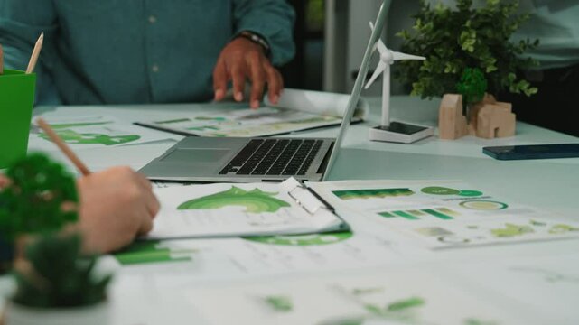 A vibrant business meeting scene showcasing professionals discussing green project plans, complete with laptops, plants, and charts, emphasizing teamwork and innovation. SACTR
