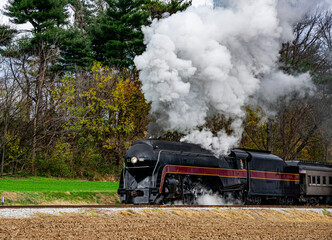 A vintage steam locomotive travels along countryside tracks, releasing clouds of black smoke into the crisp autumn air. Colorful trees provide a backdrop as the train moves forward, evoking nostalgia.