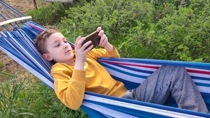Student resting during summer holidays, boy lying in hammock and looking at smartphone, online learning, outdoor recreation during summer holidays