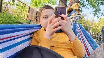 Student resting during summer holidays, boy lying in hammock and looking at smartphone, online learning, outdoor recreation during summer holidays