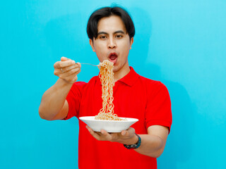 Portrait of an Asian man wearing a red polo shirt, looking excitedly at a plate of fried noodles, with a happy expression on an isolated blue background.