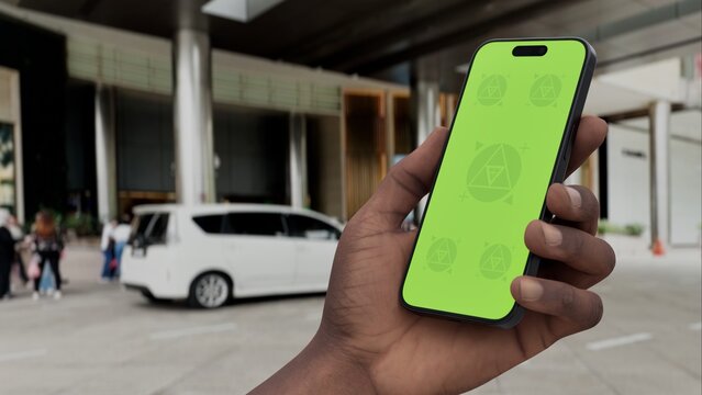 GREEN SCREEN CHROMA KEY Smartphone with a green screen held in a hand near a hotel entrance, featuring a parked car and modern urban surroundings