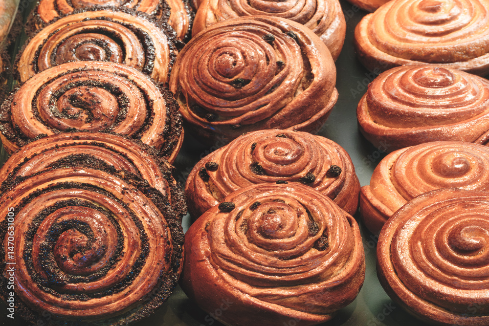 Aufkleber A row of cinnamon rolls are displayed in a bakery. The rolls are all different sizes and have different toppings. bakery is likely busy and popular, as there are many rolls on display.
