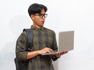 A handsome Asian male student carrying a backpack and holding a laptop, showing a funny confused...