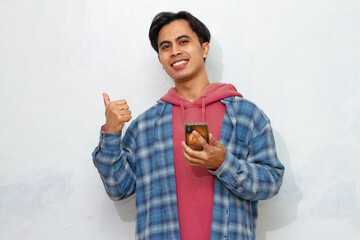 Smiling Young Asian man pointing to the side with a thumb to advertise while holding a smartphone.