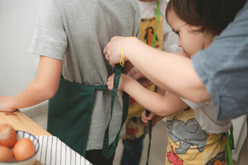 Toddler tying apron on his brother with mother's help in kitchen
