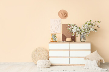 Vase with flowering branches, paintings and hat on chest of drawers in room interior