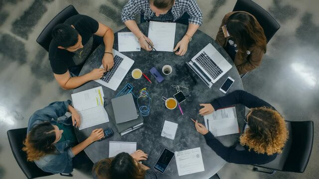 Overhead shot capturing a dynamic team working on laptops, notebooks, and documents at a round table in a modern office. Coffee cups, pens, and smartphones scattered among them.