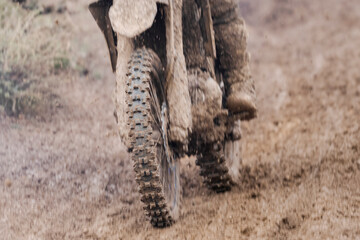  Motocross bike's front wheel and rider's boots, both heavily caked in mud, capturing the gritty intensity of an off-road race. The rugged terrain and tire tread are prominently featured, emphasizing
