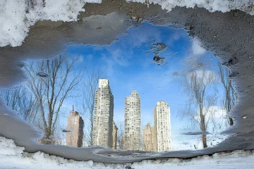 Cityscape reflected in a puddle, winter scene.  A puddle, filled with water, reflects a winter cityscape with tall buildings and bare trees. Snow and ice surround the puddle