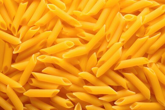Uncooked yellow penne pasta scattered on surface. Close-up view of raw Italian noodles. Healthy carbohydrate ingredient for Mediterranean cuisine. World Pasta Day concept.