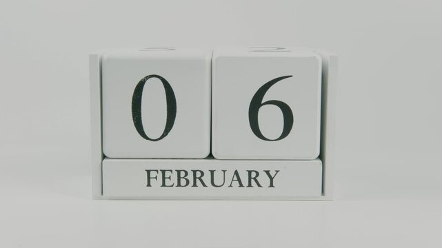 A hand flips a wooden calendar from February 6th to February 7th on a white background, marking the passing of time and the start of a new day