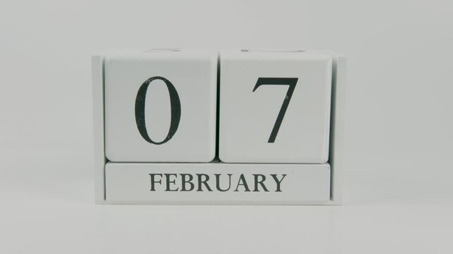 A hand flips a wooden calendar from February 7th to February 8th on a white background, marking the passing of time and the start of a new day