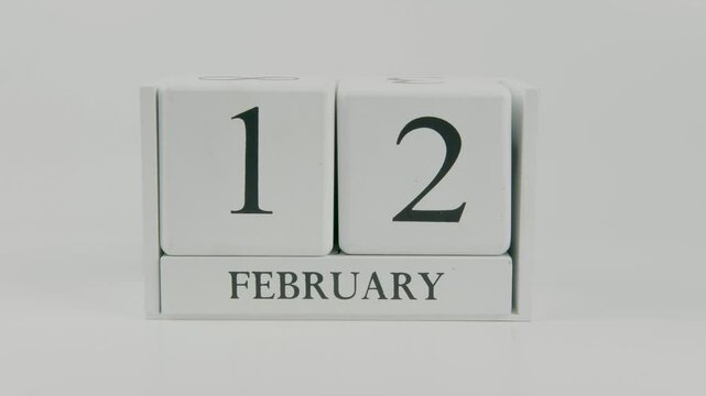 A hand flips a wooden calendar from February 12th to February 13th on a white background, marking the passing of time and the start of a new day