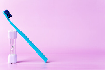 Blue toothbrush rests against a small hourglass timer on a pastel pink background, emphasizing the importance of timed dental hygiene routines