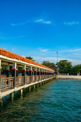 The long bridge supported by the pillar pillar structure and has a fence on both sides and a red roof that stretches over sea water and at the end of the sandy beach area bridge.