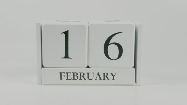 A hand flips a wooden calendar from February 16th to February 17th on a white background, marking the passing of time and the start of a new day