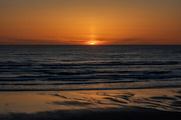 Fiery sunset at the Pacific Ocean captured from the Oregon coast. Vibrant orange sky reflecting over dark waves and wet sand. Peaceful coastal view with light textures and ocean horizon