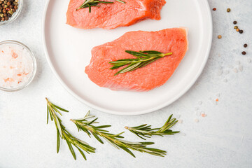 Two raw veal steaks with rosemary on white plate surrounded by cooking spices and vegetables