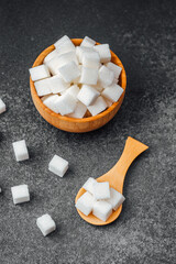 Cubes of sugar in wooden bowl and on spoon on dark stone background