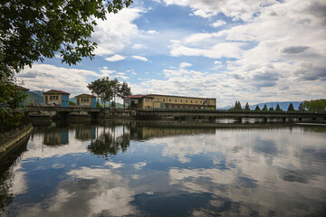 Obraz premium Hydroelectric power plant buildings and bridge reflected on the surface of a calm river under a cloudy sky