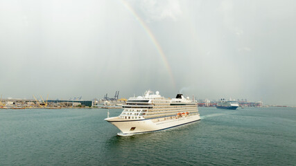 A luxury cruise ship glides through calm waters near a bustling industrial port, with a faint...