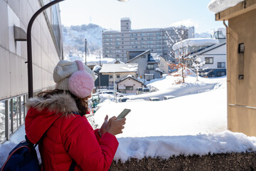 Smiling woman in red winter coat and pink earmuffs using smartphone in a snowy city street. Bright sunny day, cheerful travel moment, and winter lifestyle in an urban environment.