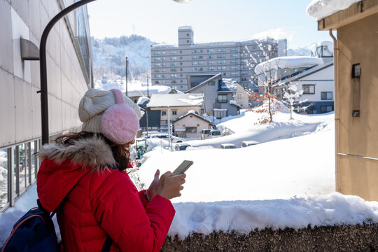 Smiling woman in red winter coat and pink earmuffs using smartphone in a snowy city street. Bright sunny day, cheerful travel moment, and winter lifestyle in an urban environment. - Powered by Adobe