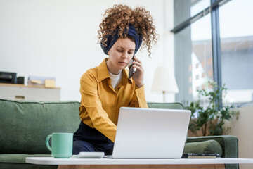 Confident Russian businesswoman with curly hair sits on modern office sofa, writing in notebook. plans strategies and documents ideas with thoughtful intent and professional poise.