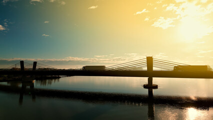 A truck crosses a bridge over a river at sunset, symbolizing the continuous flow of cargo and...