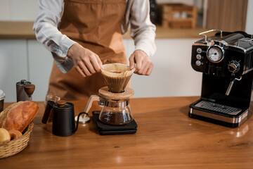 A young woman with curly hair and a headband, wearing a brown apron, prepares pour-over coffee on a wooden countertop in a bright kitchen. Focused and calm, she enjoys her morning routine.
